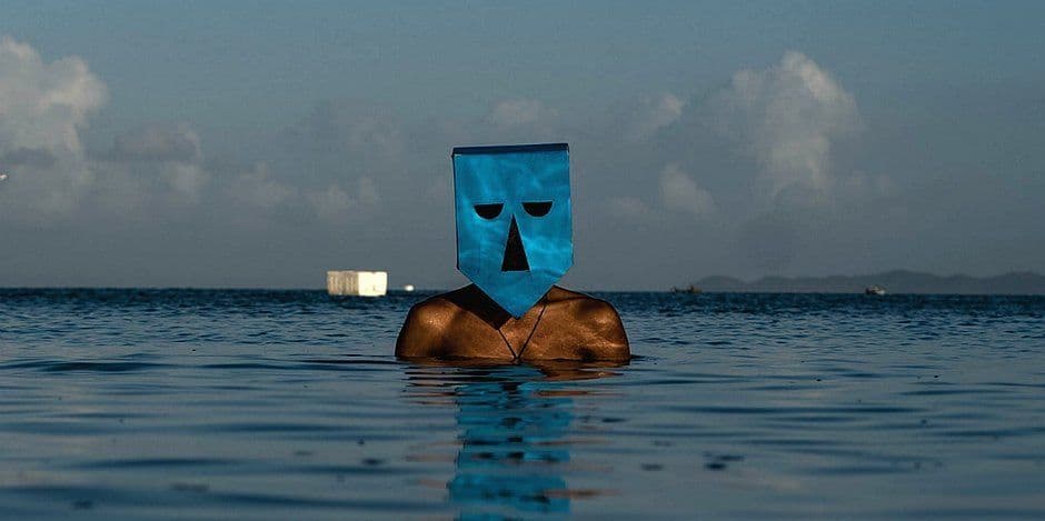 Pessoa com o torso parcialmente submerso no mar usa uma máscara azul geométrica, com nuvens e montanhas ao fundo.