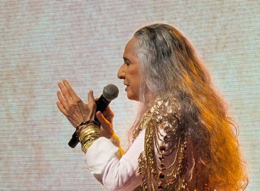 Maria Bethânia de perfil, segurando um microfone, com roupa branca e adornos dourados.