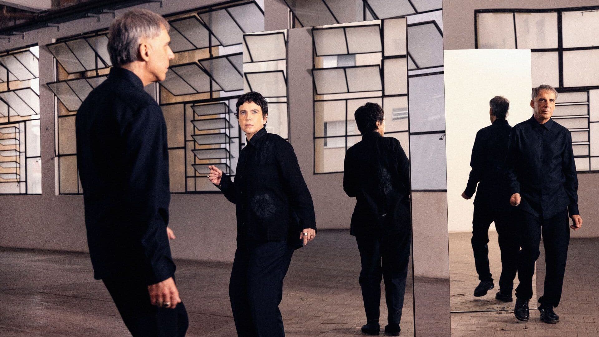 Adriana Calcanhotto e Arnaldo Antunes vestidos de preto, diante de espelhos que refletem suas imagens.