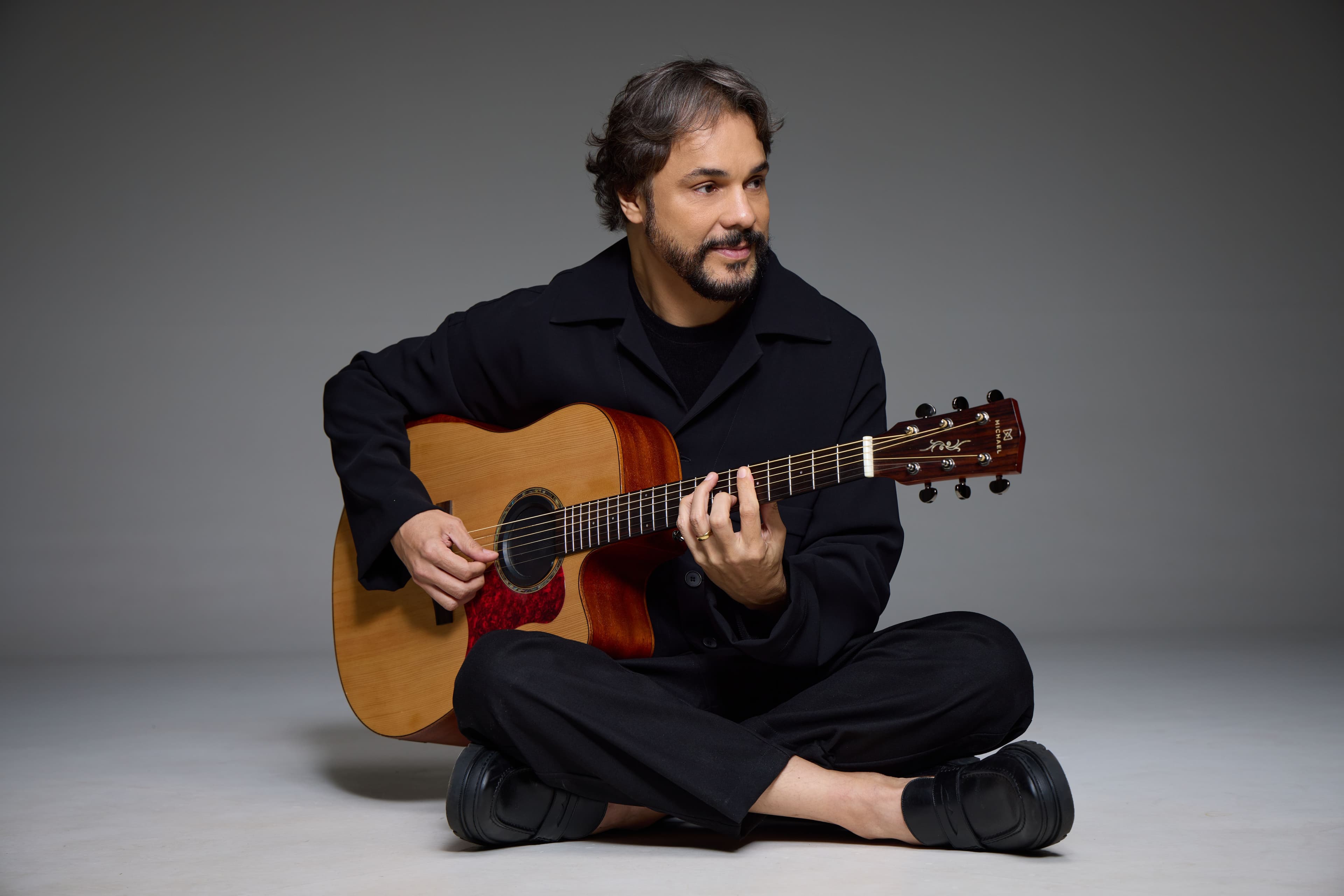 Rodrigo Borges sentado de pernas cruzadas, segurando um violão, com roupa preta e fundo cinza.