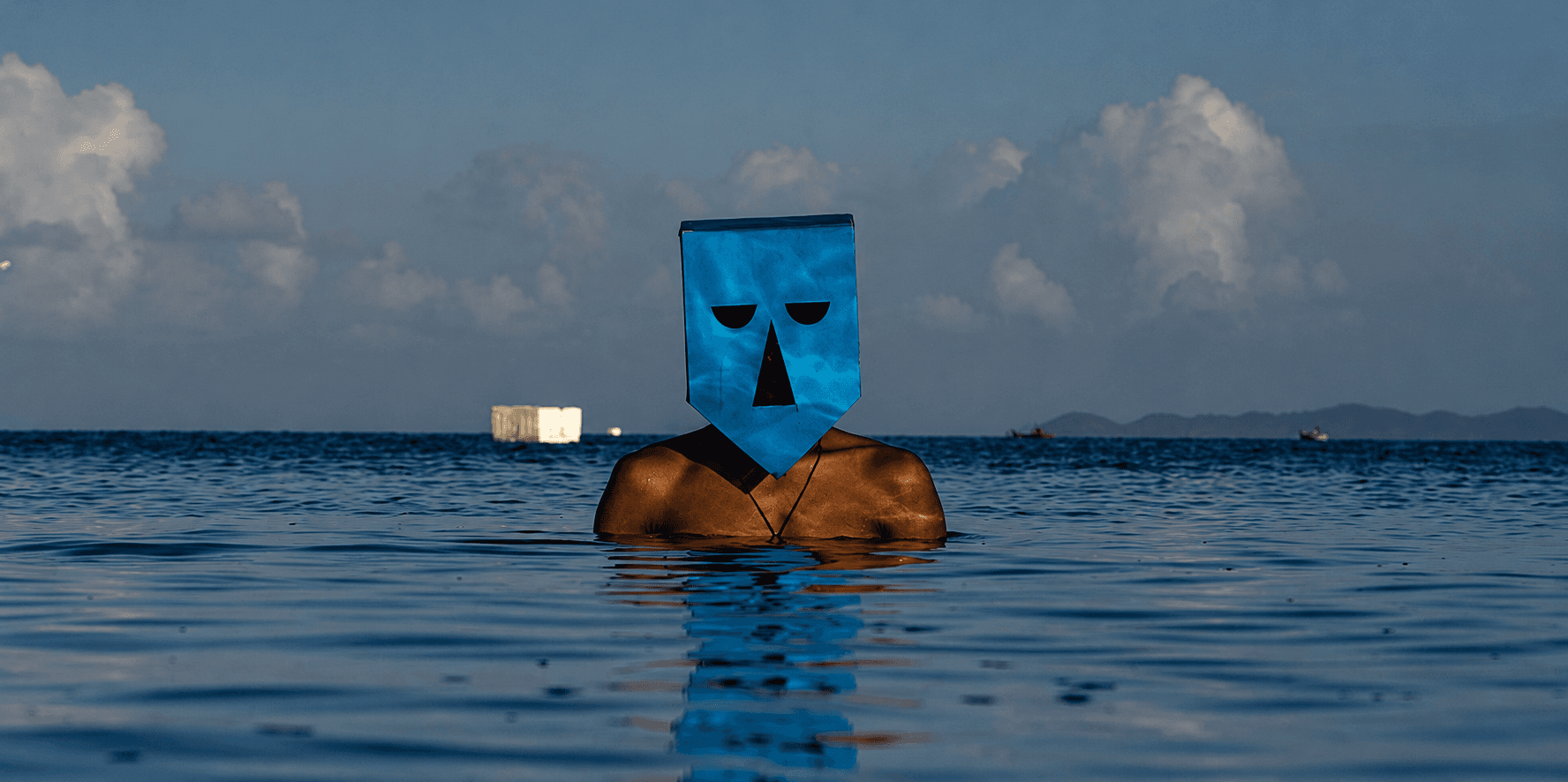 Pessoa com o torso parcialmente submerso no mar usa uma máscara azul geométrica, com nuvens e montanhas ao fundo.