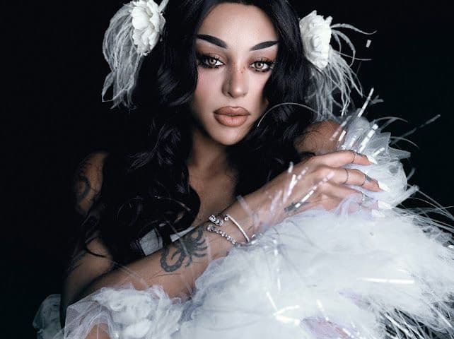 Pabllo Vittar com cabelos pretos longos, adornos brancos e roupa com plumas, diante de um fundo escuro.